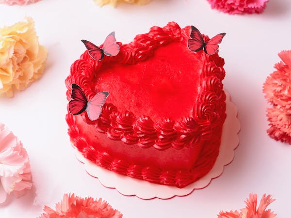 A vibrant red heart-shaped cake adorned with butterfly toppers surrounded by colorful carnations on a white background.
