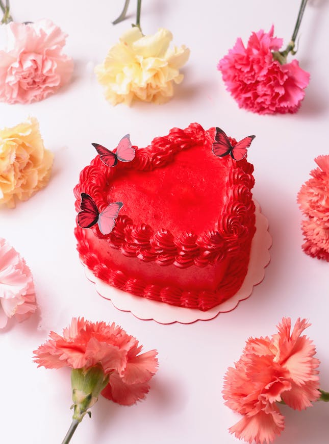 Foto van Sergio Arreola A vibrant red heart-shaped cake adorned with butterfly toppers surrounded by colorful carnations on a white background.