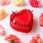 A vibrant red heart-shaped cake adorned with butterfly toppers surrounded by colorful carnations on a white background.